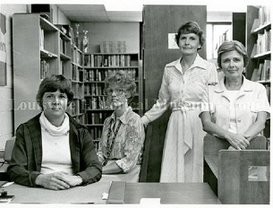 Four women posed by a reference desk with bookshelves