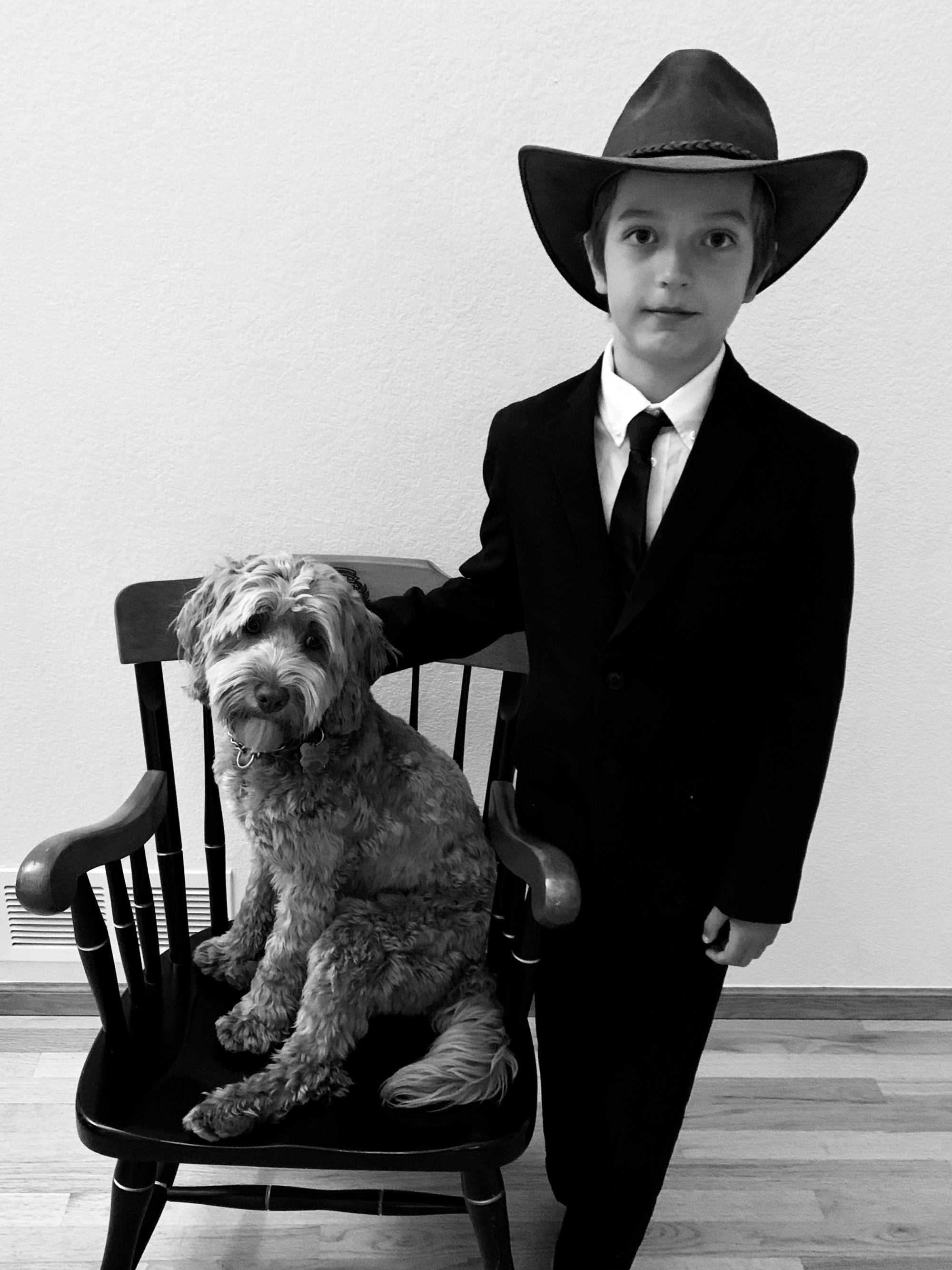 Boy in suit with large hat posing with small dog sitting on a chair.