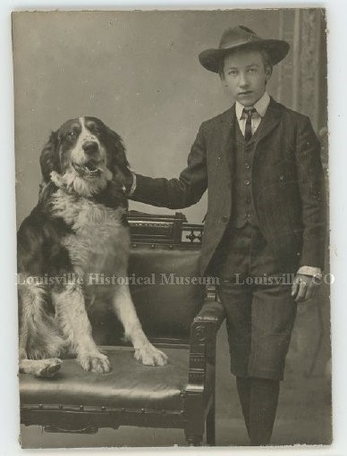 Old photo of boy in suit and hat posing with a large dog sitting on a chair.