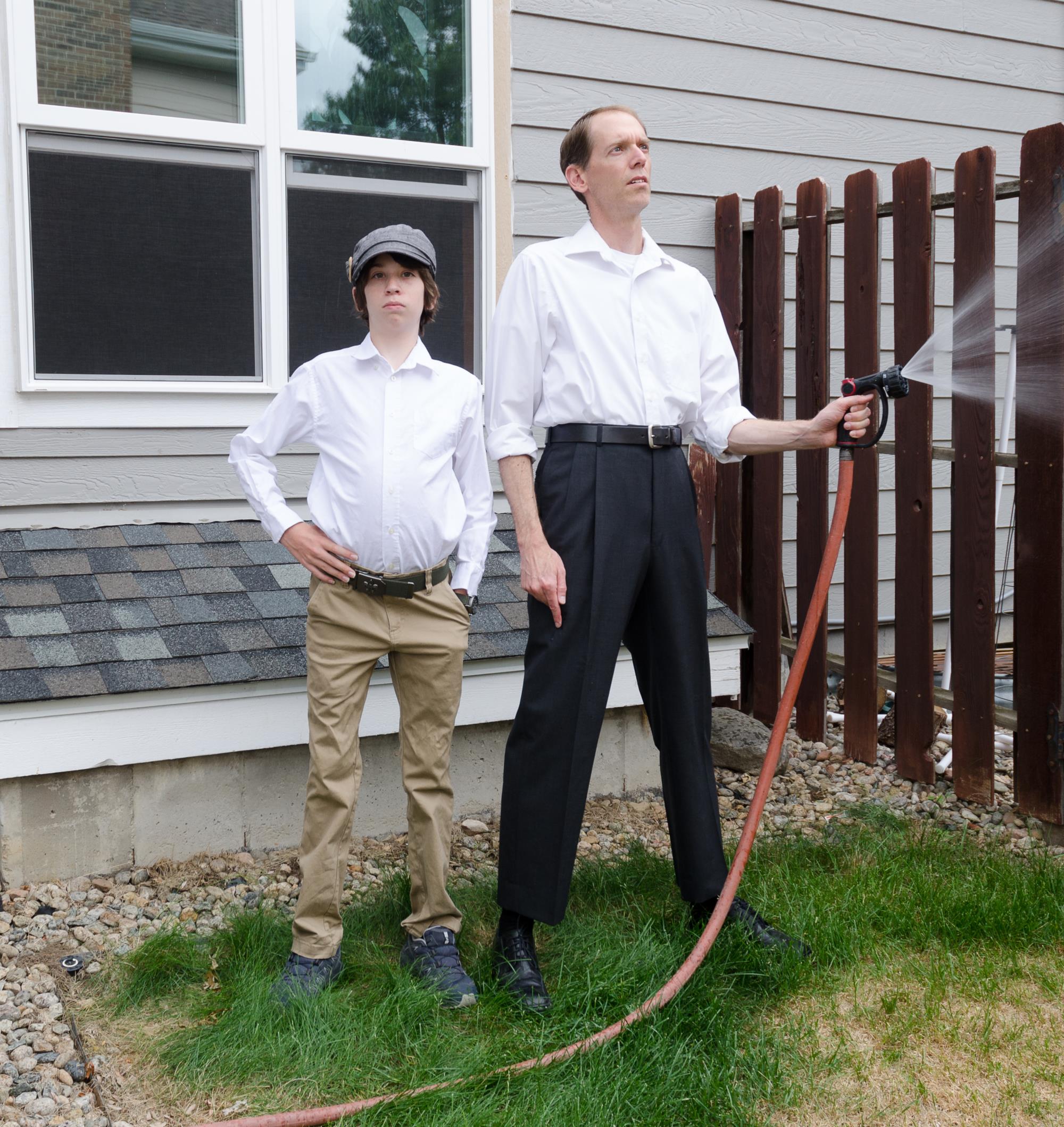 Man watering lawn with hose and boy standing with hands on hips.