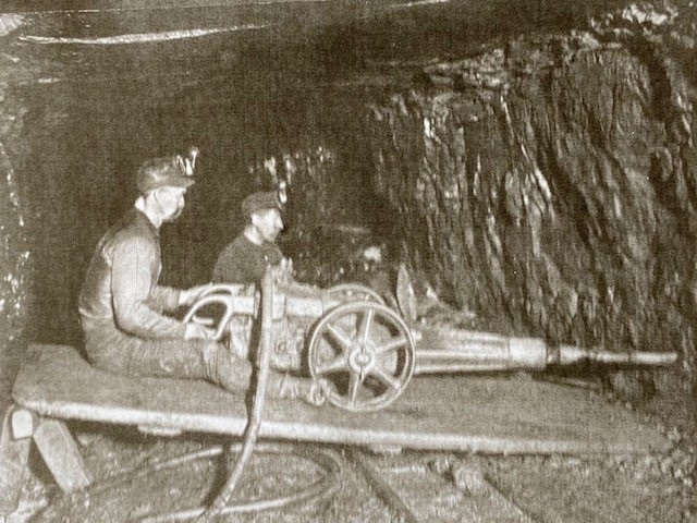 Old photo of two miners straddling a punching machine in a coal mine.