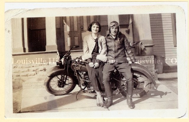 Man in leather jacket and helmet and woman leaning against a motorcycle.