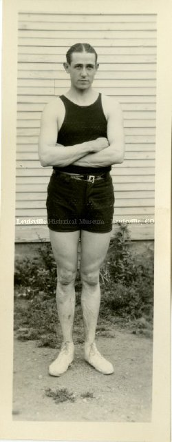 Old photo of man in tank top and shorts standing with arms crossed.