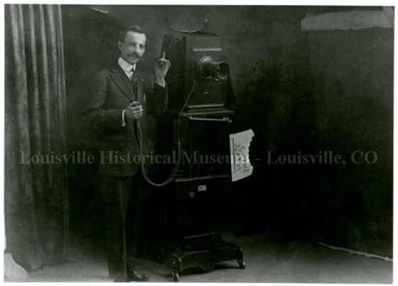 Man posing with large camera on stand with sign reading &quot;smile please&quot;.