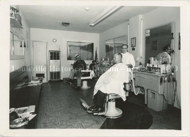 Interior of barbershop with barber and two men sitting in chairs.