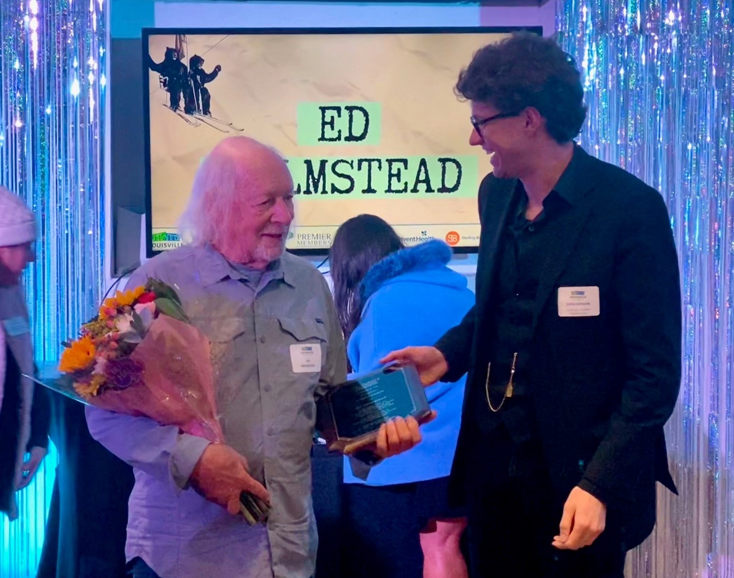 A man holding flowers with his name projected behind him, "Ed Helmstead" receiving a plaque from a man in a suit