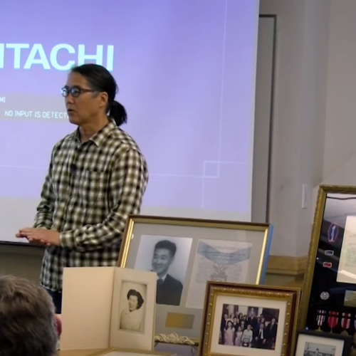 A Japanese American man stands in front of family portraits and framed military honors