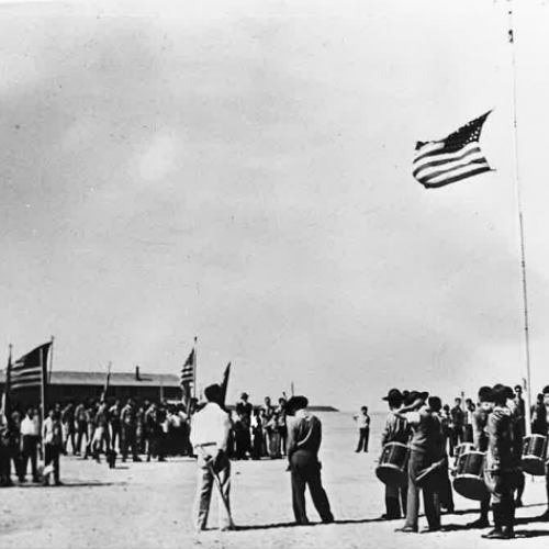 black and white photo of men in an interment camp with the Unites States flag flying above them